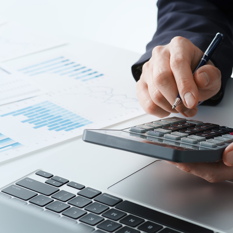 Main d'un professionnel en costume tenant un stylo au-dessus d'une calculatrice, travaillant avec un ordinateur portable et des graphiques financiers.